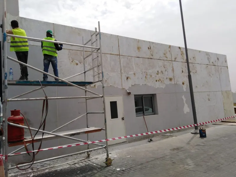 Stained and peeling external wall panels with rust marks at Porsche service centre Sharjah, workers on mobile scaffold